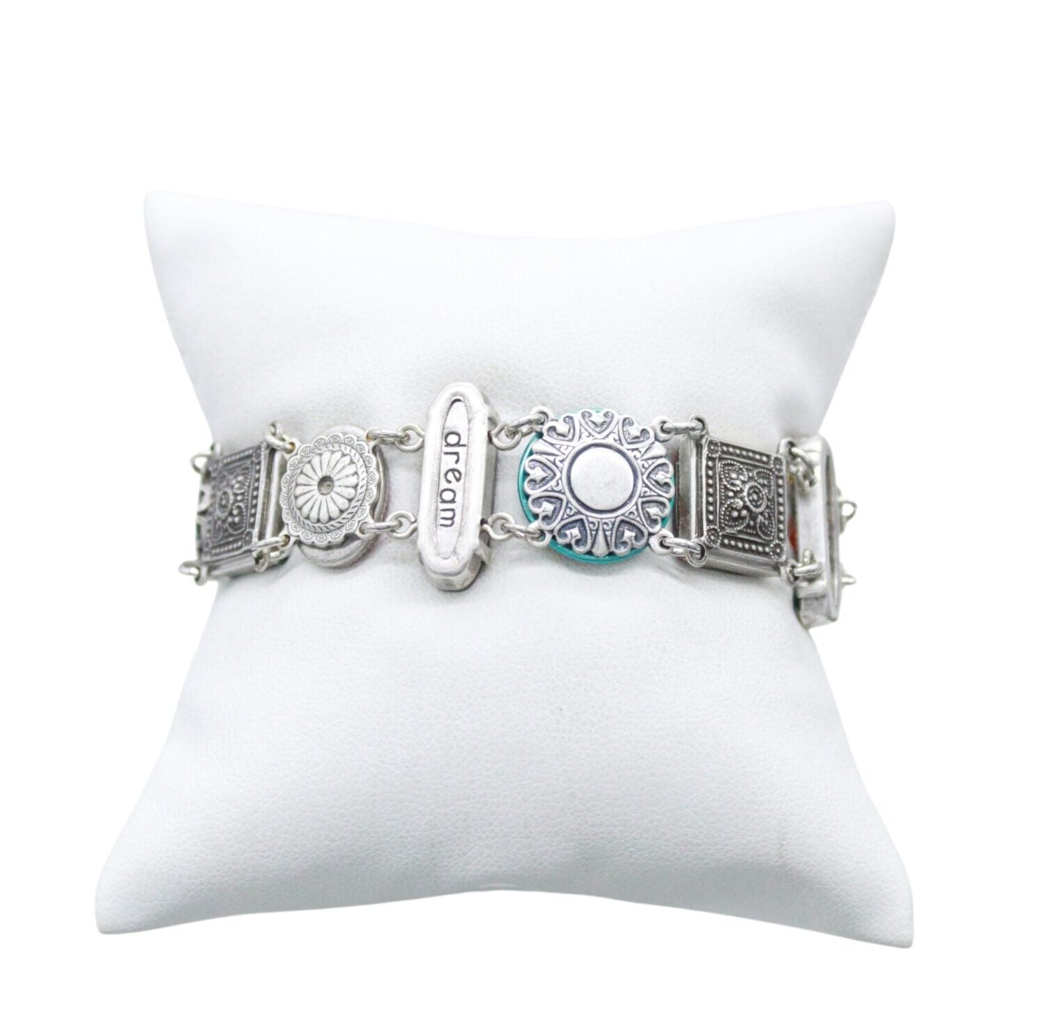 The Firefly La Dolce Vita Multi-Color Bracelet boasts a stunning silver tone and fair-trade craftsmanship. Its floral-patterned links and oval plate engraved with dream are beautifully displayed on a white cushioned stand.