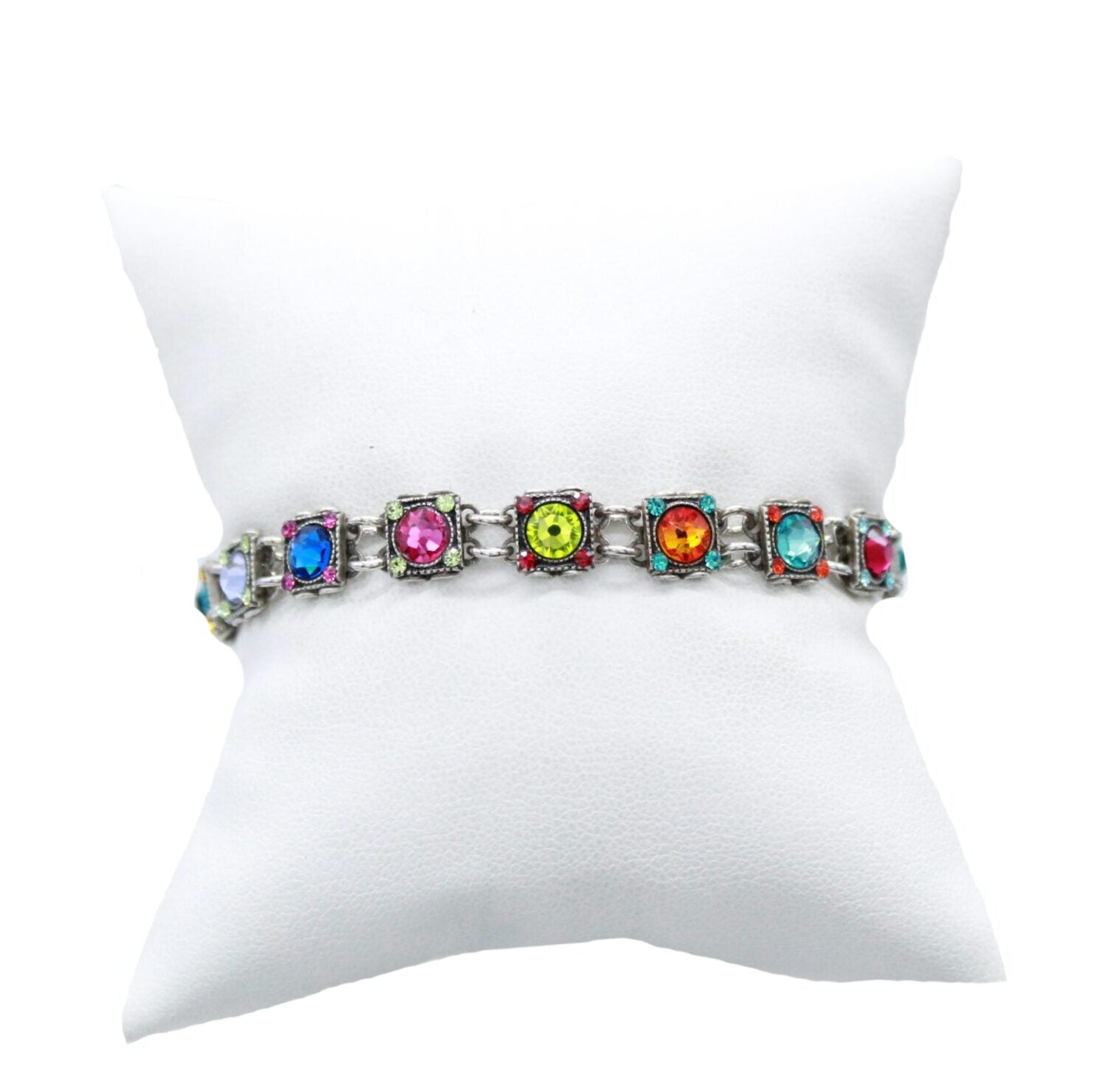 The Thin Rainbow Square Bracelet by Firefly Jewelry, handmade in Guatemala, adorns a white pillow. This fair-trade piece showcases multicolored gemstones like red, blue, green, and pink beautifully set on a silver bracelet.