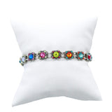 The Thin Rainbow Square Bracelet by Firefly Jewelry, handmade in Guatemala, adorns a white pillow. This fair-trade piece showcases multicolored gemstones like red, blue, green, and pink beautifully set on a silver bracelet.