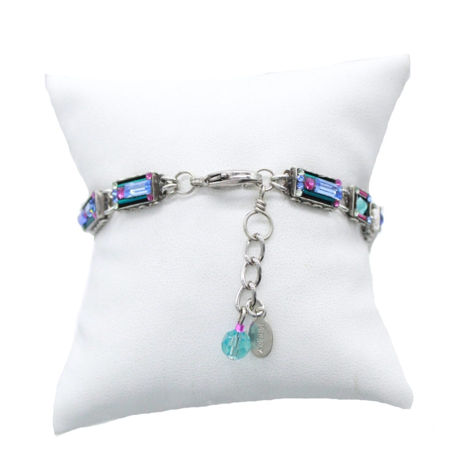 The Architectural Light Turquoise Bracelet by Firefly Jewelry, handmade in Guatemala, showcases multicolored rectangular stones on a silver band, with a dangling blue bead and metal tag near the clasp, displayed on a white cushion.