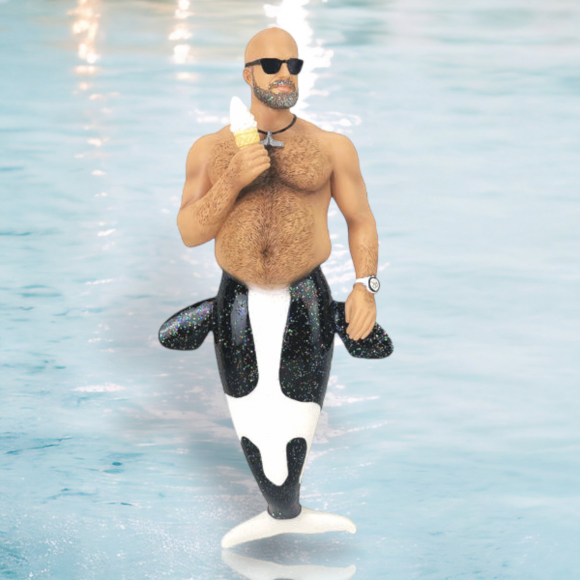 A Beached Whale/Black Tail Ornament by December Diamonds features a shirtless merman with an orca tail. He stands in the water, wearing sunglasses and holding an ice cream cone, with a watch on his wrist accentuating his muscular build against a light blue watery background.
