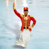 The Nut Cracker Ornament by December Diamonds features a hand-painted merman in a red and gold marching band uniform, with a detailed fish-scale tail. Against a watery blue background, this enchanting piece is perfect for holiday decor.