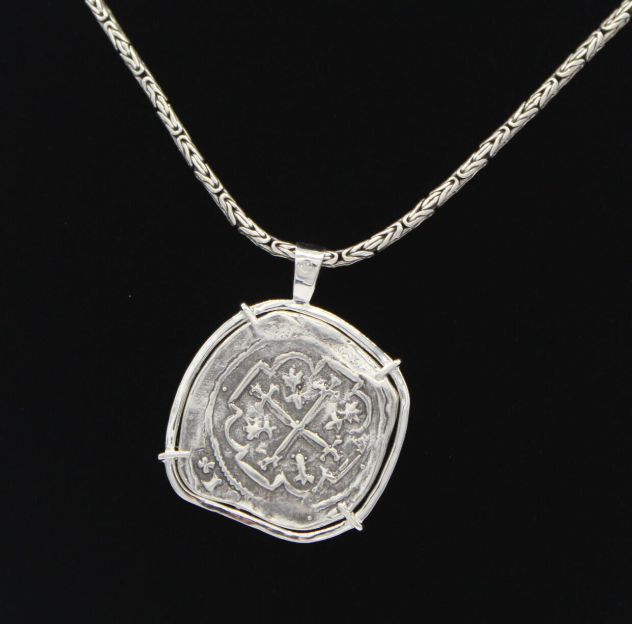 The Treasure Shoppe Inc.s Curved 8 Reale Coin- Atocha Shipwreck Coins features an engraved silver pendant, reminiscent of an old Atocha coin, framed in a sterling silver bezel, hanging from a woven chain against a plain black background.