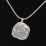 The Treasure Shoppe Inc.s Curved 8 Reale Coin- Atocha Shipwreck Coins features an engraved silver pendant, reminiscent of an old Atocha coin, framed in a sterling silver bezel, hanging from a woven chain against a plain black background.