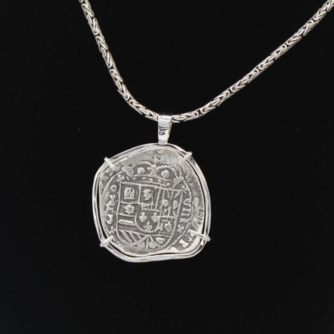 The Treasure Shoppe Inc.s Curved 8 Reale Coin Pendant, inspired by Atocha Shipwreck coins, features an ornate silver pendant set in a sterling silver bezel. The woven chain and black background enrich its intricate design.