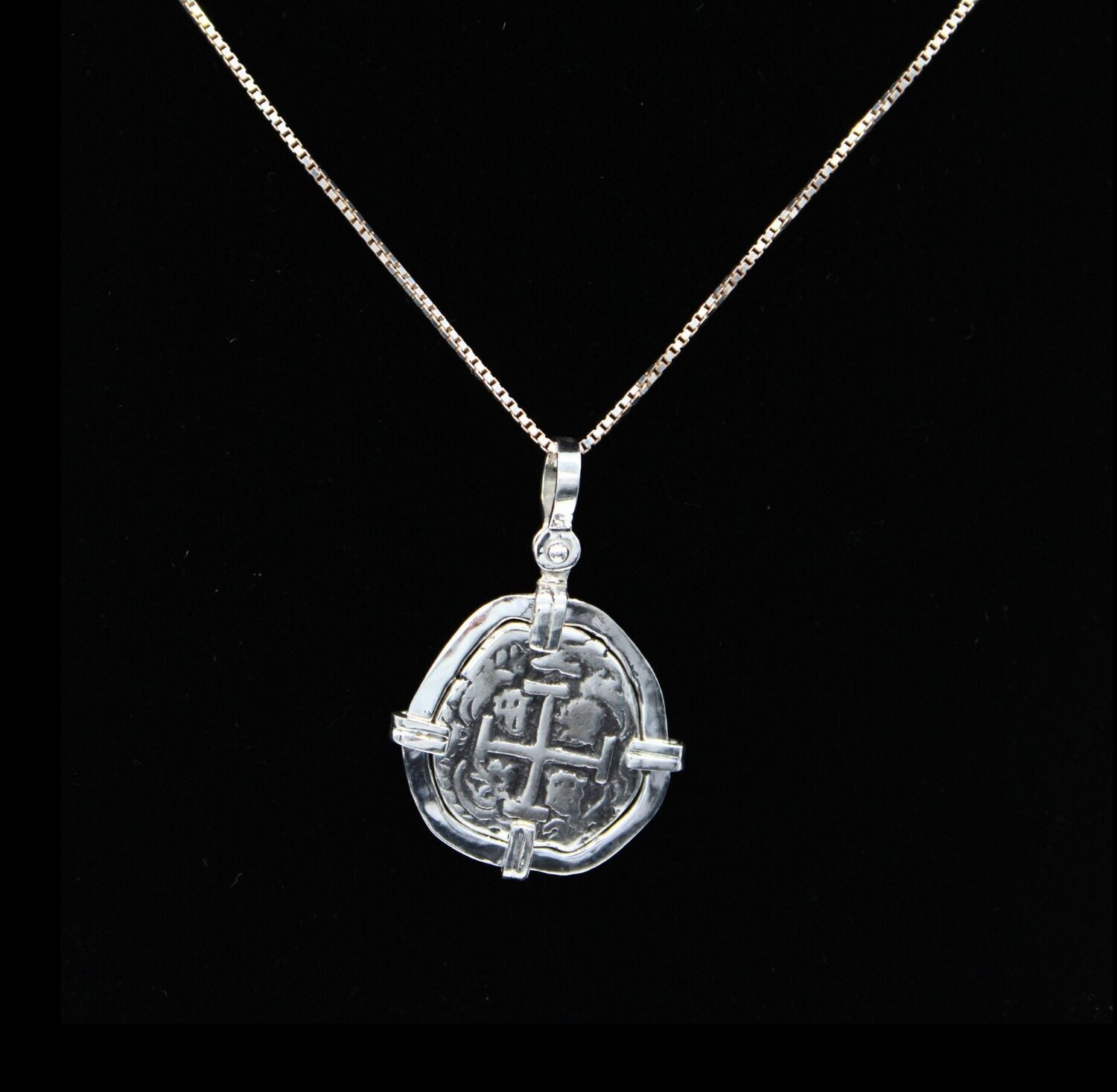 Set against a black backdrop, The Treasure Shoppe Inc.s sterling silver Atocha 302 Coin pendant captures the historical essence of Atocha shipwreck coins. Its worn engravings feature a central cross with indistinct details, elegantly hanging from the thin silver chain.