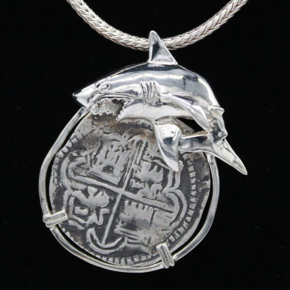 The Treasure Shoppe Inc.s 8 Reale Shark Coin Pendant, crafted in sterling silver, features a shark motif over a round Atocha shipwreck coin with cross-like symbols. The intricate metalwork stands out against its black background and comes with a braided chain for wearing.