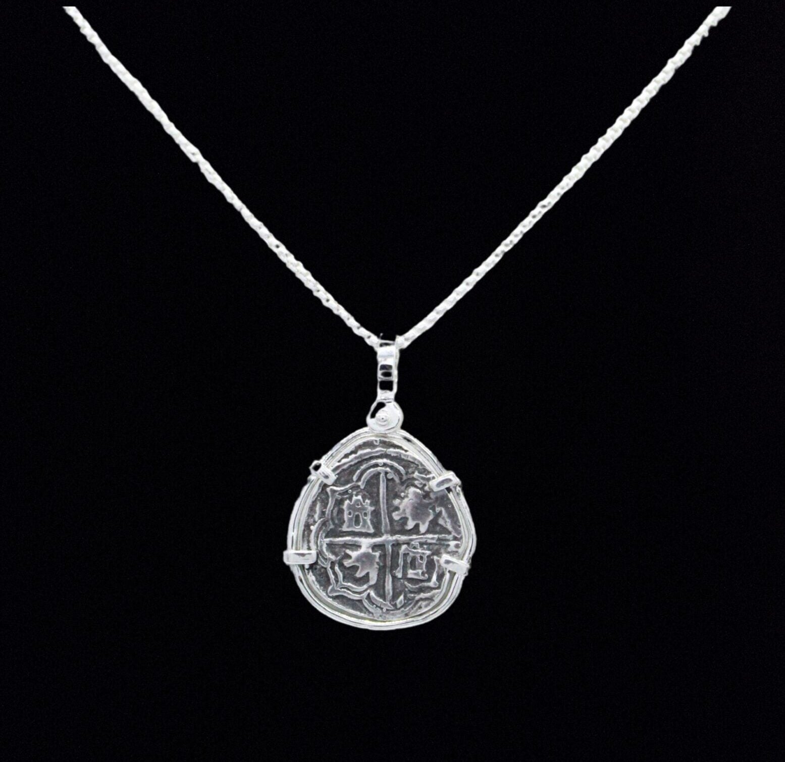The Treasure Shoppe Inc.s Reale/Oblong Silver Frame Coin pendant features a rustic, coin-like design inspired by historic Nuestra Señora de Atocha coins. Its intricate patterns create an antique feel against a black backdrop, making it stand out on any fine silver chain.