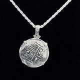 The Treasure Shoppe Inc.s 500 Museum Replica Coin, inspired by Atocha Shipwreck Coins, showcases a silver pendant with worn engravings and an elegant wire setting. It stands out against a black background with its antique appearance enhanced by a sterling silver bezel.
