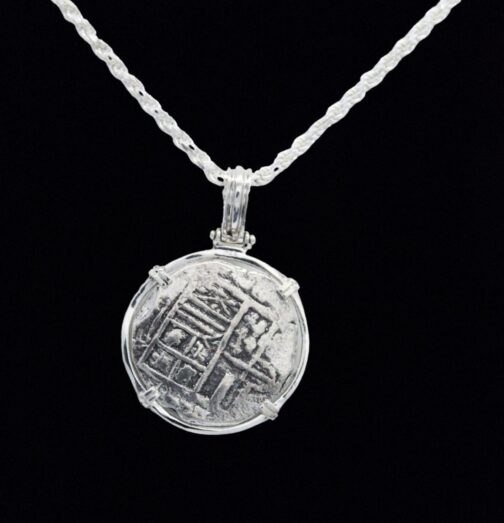 The Treasure Shoppe Inc.s 500 Museum Replica Coin, inspired by Atocha Shipwreck Coins, showcases a silver pendant with worn engravings and an elegant wire setting. It stands out against a black background with its antique appearance enhanced by a sterling silver bezel.
