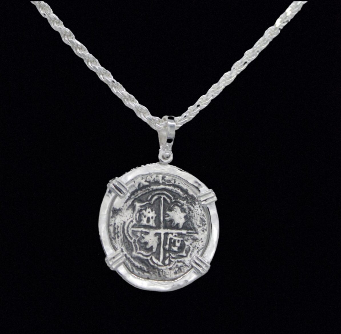 The 502 Coin pendant from The Treasure Shoppe Inc. is a sterling silver piece resembling an antique Atocha shipwreck coin, featuring a cross and shield design. Its historic charm shines against a black background, hanging from a textured chain.
