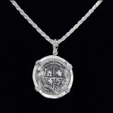 The 502 Coin pendant from The Treasure Shoppe Inc. is a sterling silver piece resembling an antique Atocha shipwreck coin, featuring a cross and shield design. Its historic charm shines against a black background, hanging from a textured chain.