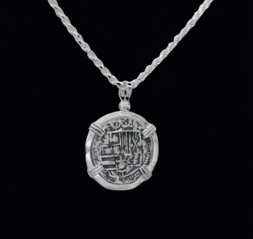 The Treasure Shoppe Inc.s 502 Coin pendant, reminiscent of Atocha shipwreck coins, features intricate ancient-style engravings on sterling silver. The design is highlighted by a twisted chain and black background to showcase its historic charm.
