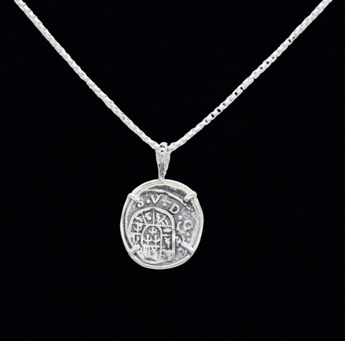 The Treasure Shoppe Inc.s 1 Reale Coin- Atocha Shipwreck Pendant is a historic piece featuring intricate markings on a sterling silver round pendant with a black background, suspended from a thin, textured chain.
