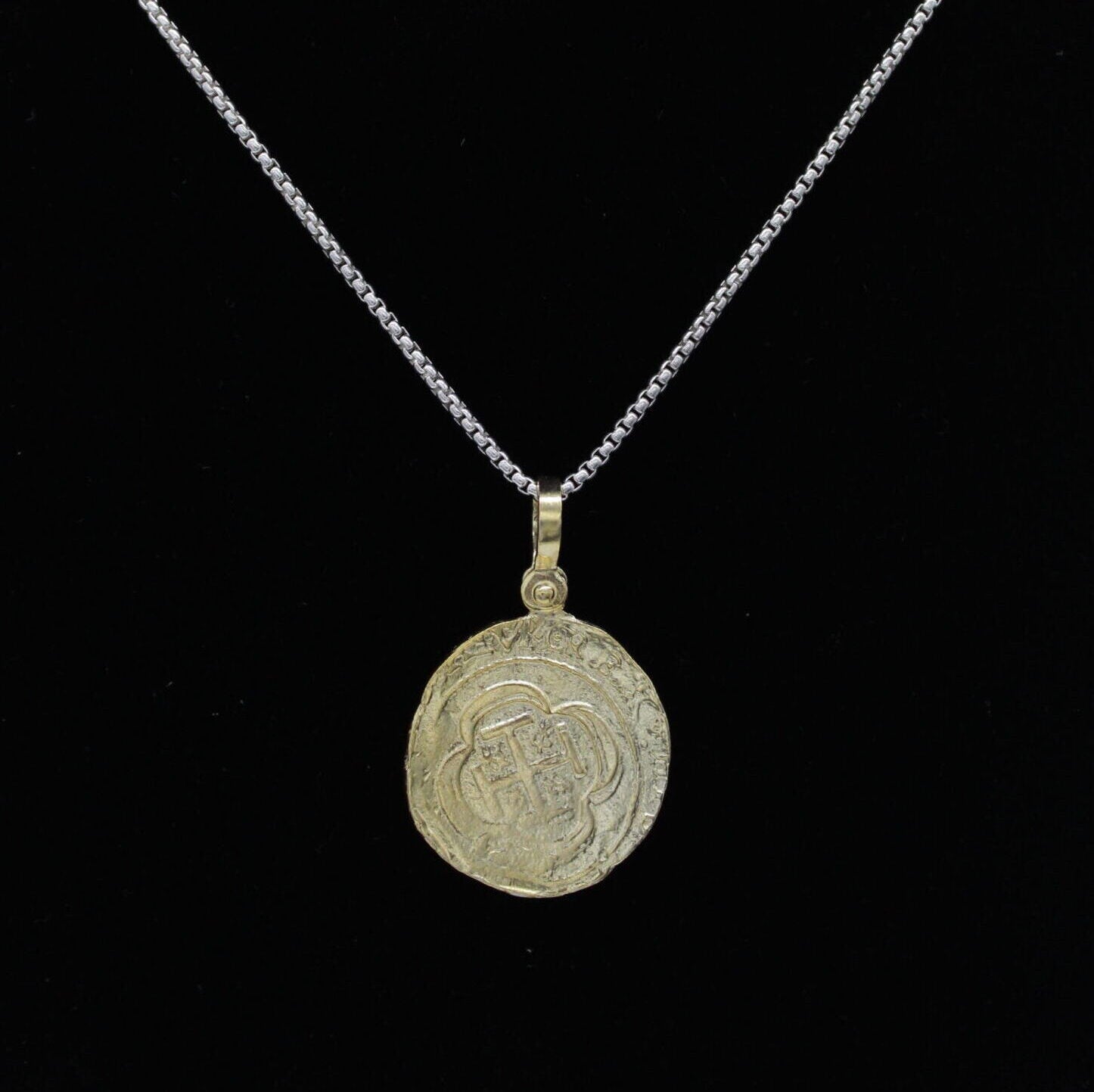 The Treasure Shoppe Inc.s Gold Plated Round Escudo Coin pendant mimics Atocha shipwreck coins with its intricate patterns and symbols. The weathered coin-like charm stands out against a solid black background, highlighting its unique design.