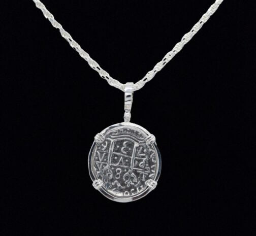 The Treasure Shoppe Inc.s 8 Reale Triple Prong Coin pendant showcases an ornate, engraved circular design with symbols and numbers, evoking historic charm. The sterling silver piece hangs elegantly on a twisted chain against a black background.