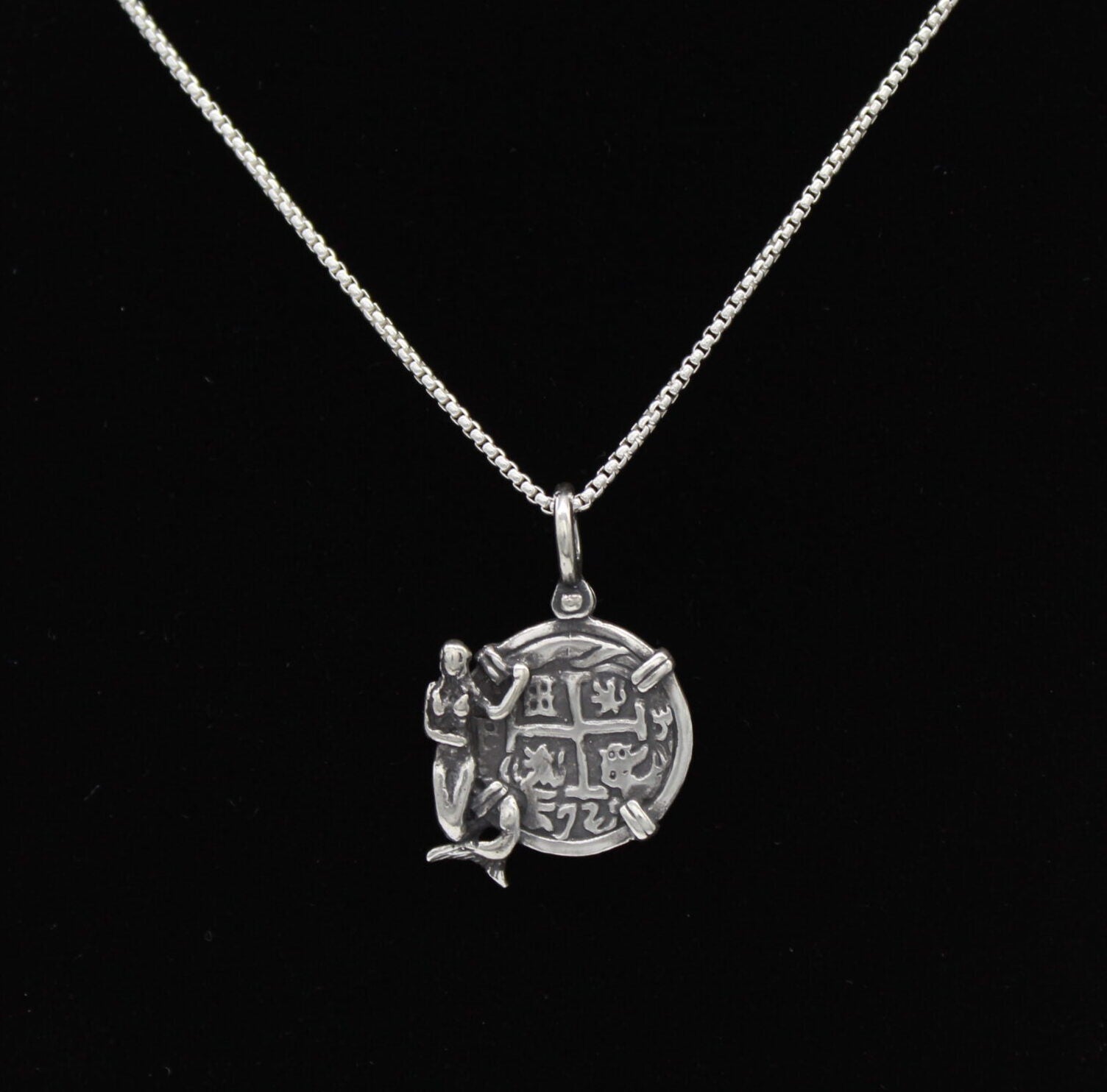 The Treasure Shoppe Inc.s Medium Mermaid Coin features an engraved round sterling silver bezel pendant with symbols and a detailed figure on the left, reminiscent of shipwreck coins. The pendant is displayed against a black background without a chain.