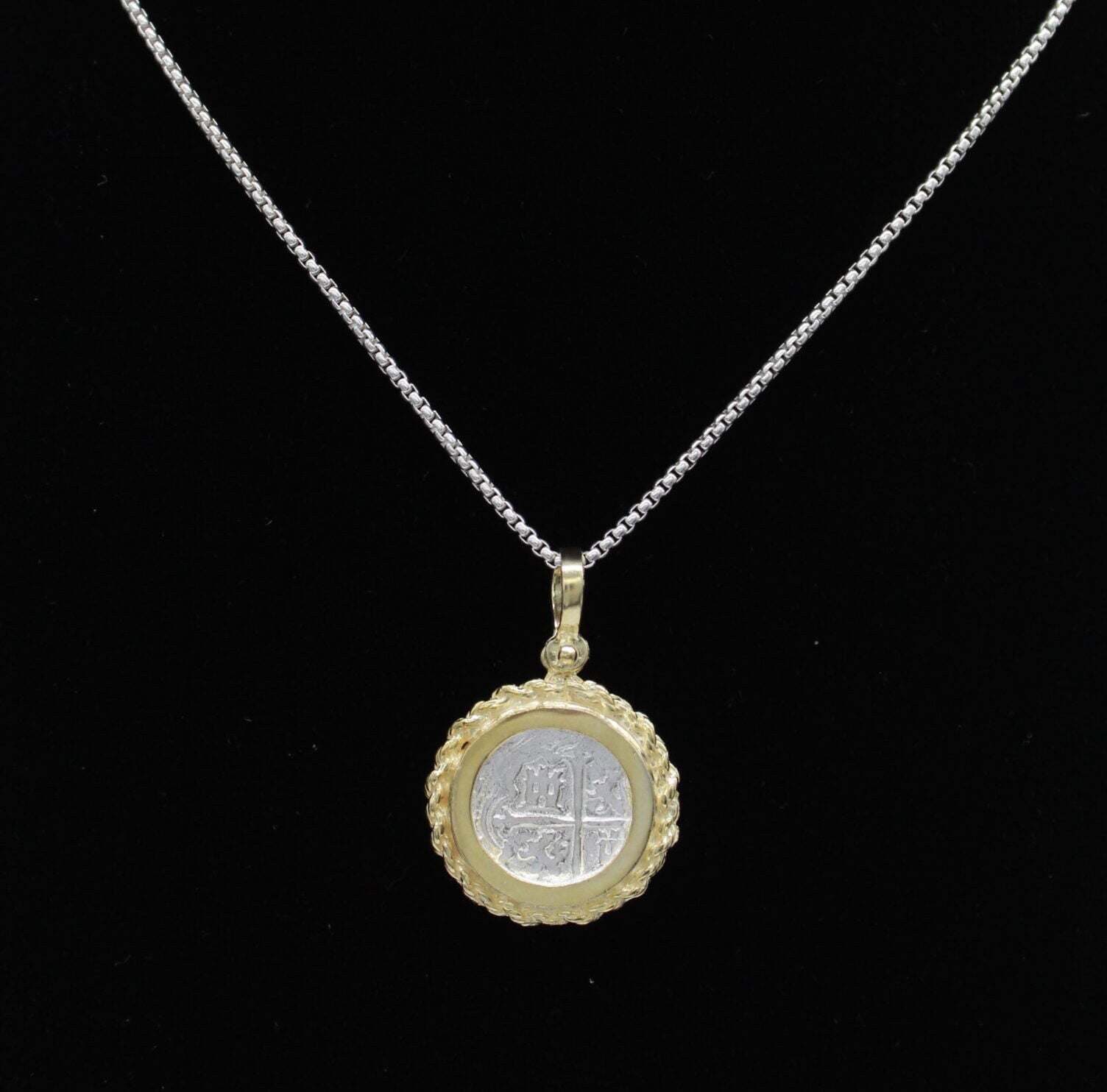 A silver necklace features a round coin pendant with an ornate gold-plated rope bezel and embossed center design, set against a black background, from The Treasure Shoppe Inc.s Gold Plated Rope Bezel Coin - Atocha Shipwreck Coins collection.