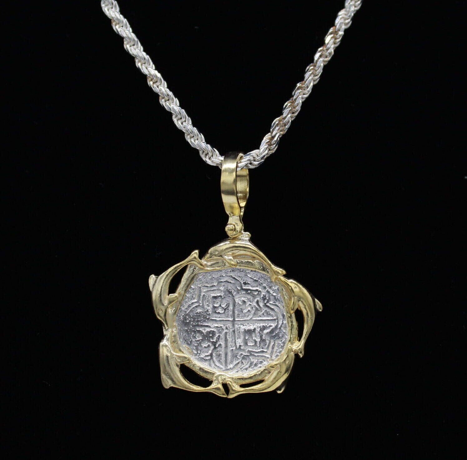 The Treasure Shoppe Inc.s Gold Plated Dolphin Frame Coin, inspired by Atocha shipwreck treasures, features a rustic silver coin pendant set in an ornate gold plated design, hanging on a thick silver rope with a solid black backdrop.