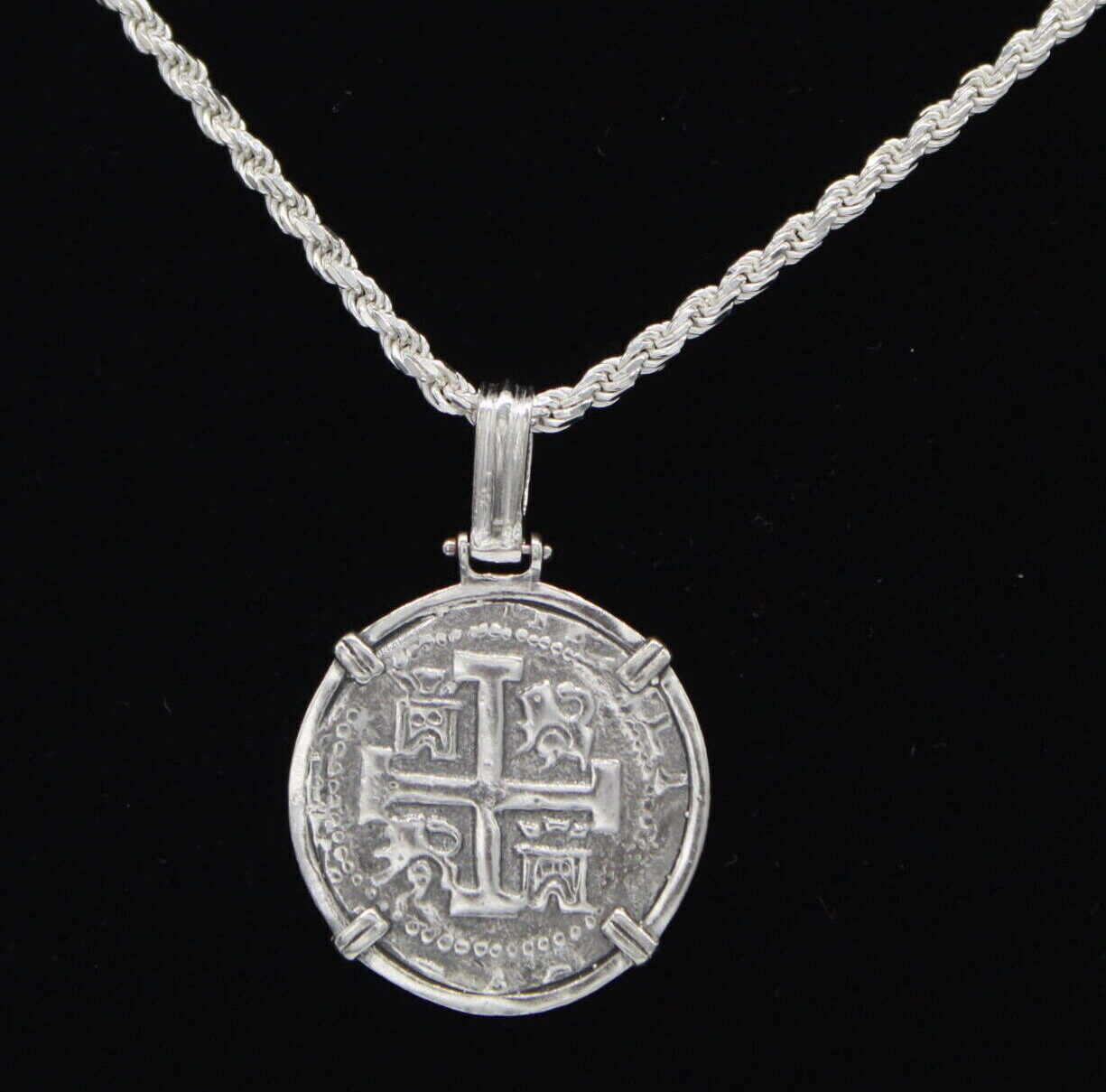 The Treasure Shoppe Inc.s 8 Reale 2 Prong Coin-Atocha Shipwreck Pendant boasts intricate designs in a simple metal frame. It dangles from a twisted sterling silver bezel chain against a black background, reminiscent of Atocha Coins.