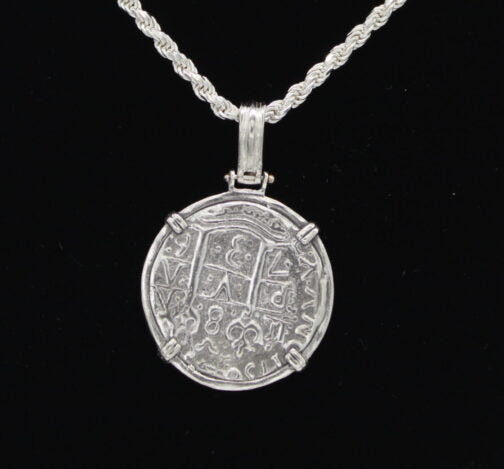 On a black backdrop, a round silver pendant from The Treasure Shoppe Inc.s 8 Reale 2 Prong Coin-Atocha Shipwreck Coins collection hangs in a twisted sterling silver bezel. Resembling Atocha Coins, its secured by four brackets and showcases intricate symbols and patterns for an antique look.