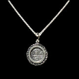 The Treasure Shoppe Inc.s Silver Rope Bezel Coin pendant features an engraved, ancient coin-like design reminiscent of Atocha shipwreck coins. Its braided silver edge and solid black background accentuate the intricate details.