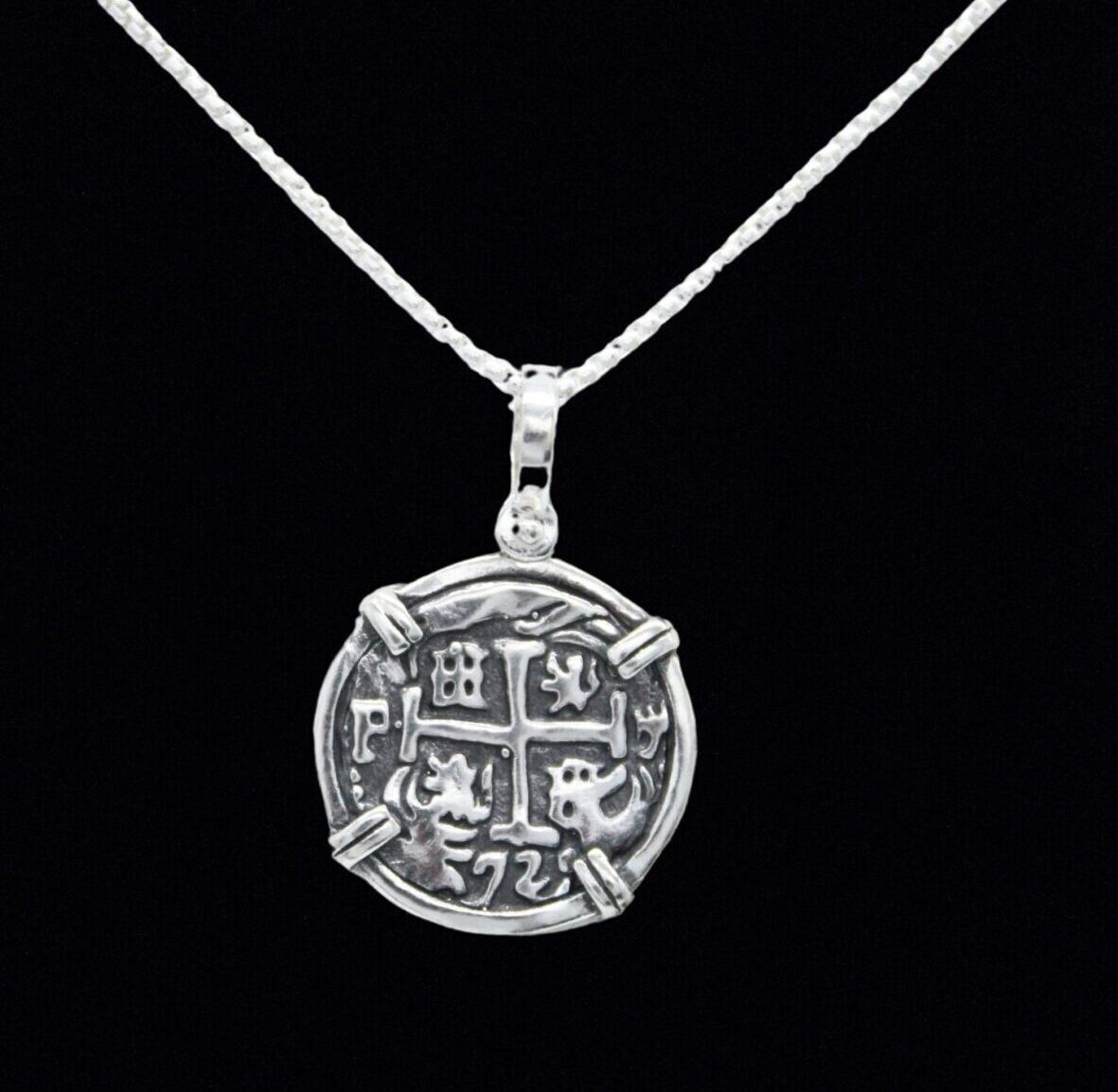 This pendant, the Silver Round Reale Coin by The Treasure Shoppe Inc., features a design inspired by an Atocha coin. Intricate symbols and numbers frame a cross, encased in a sterling silver bezel with four clips, highlighting its historic charm against a black background.
