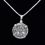 This pendant, the Silver Round Reale Coin by The Treasure Shoppe Inc., features a design inspired by an Atocha coin. Intricate symbols and numbers frame a cross, encased in a sterling silver bezel with four clips, highlighting its historic charm against a black background.