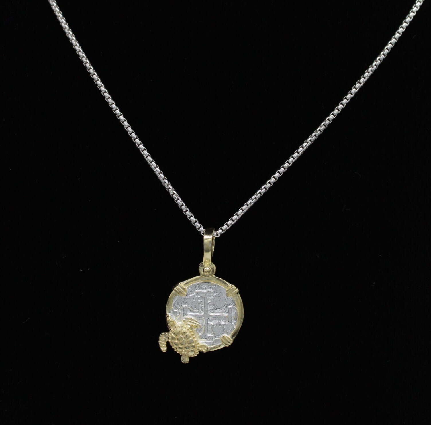 A silver necklace with a circular pendant, reminiscent of historic charm jewelry, features intricate designs and is adorned with a small gold decorative element at the bottom. The Gold Plated Small Turtle Coin from The Treasure Shoppe Inc., rests elegantly against a plain black background.
