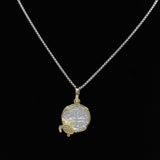 A silver necklace with a circular pendant, reminiscent of historic charm jewelry, features intricate designs and is adorned with a small gold decorative element at the bottom. The Gold Plated Small Turtle Coin from The Treasure Shoppe Inc., rests elegantly against a plain black background.
