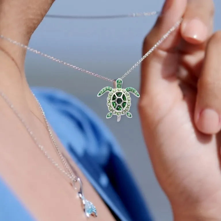 A person adjusts their Shanore Sterling Silver August Birthstone Turtle Necklace with green Swarovski crystals. They wear a blue garment against a softly blurred background.