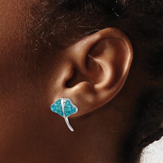 A close-up of an ear wearing Presco/Quality Gold Crystal Stingray Teal Post Earrings. The background features the persons braided hair and brown skin.