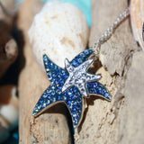 The Shanore Swarovski Crystals Dark Blue Starfish Necklace-Ocean Jewelry features a sterling silver necklace with a blue starfish pendant and a sparkling Swarovski crystal-encrusted starfish, elegantly displayed on driftwood with seashells in the backdrop.
