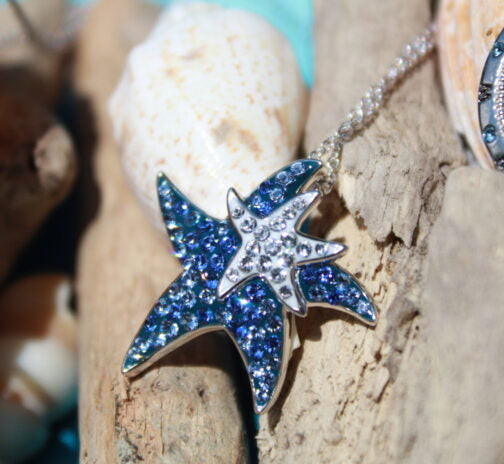 The Shanore Swarovski Crystals Dark Blue Starfish Necklace-Ocean Jewelry features a sterling silver necklace with a blue starfish pendant and a sparkling Swarovski crystal-encrusted starfish, elegantly displayed on driftwood with seashells in the backdrop.