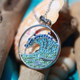 The Shanore Swarovski Crystals Blue Wave Necklace - Ocean Jewelry presents a sterling silver circle with blue and green crystals in a wave design, reminiscent of the sea, elegantly displayed against driftwood and seashells.