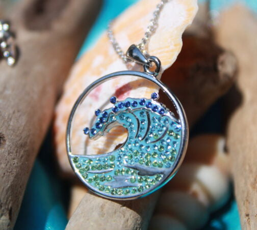 The Shanore Swarovski Crystals Blue Wave Necklace - Ocean Jewelry presents a sterling silver circle with blue and green crystals in a wave design, reminiscent of the sea, elegantly displayed against driftwood and seashells.