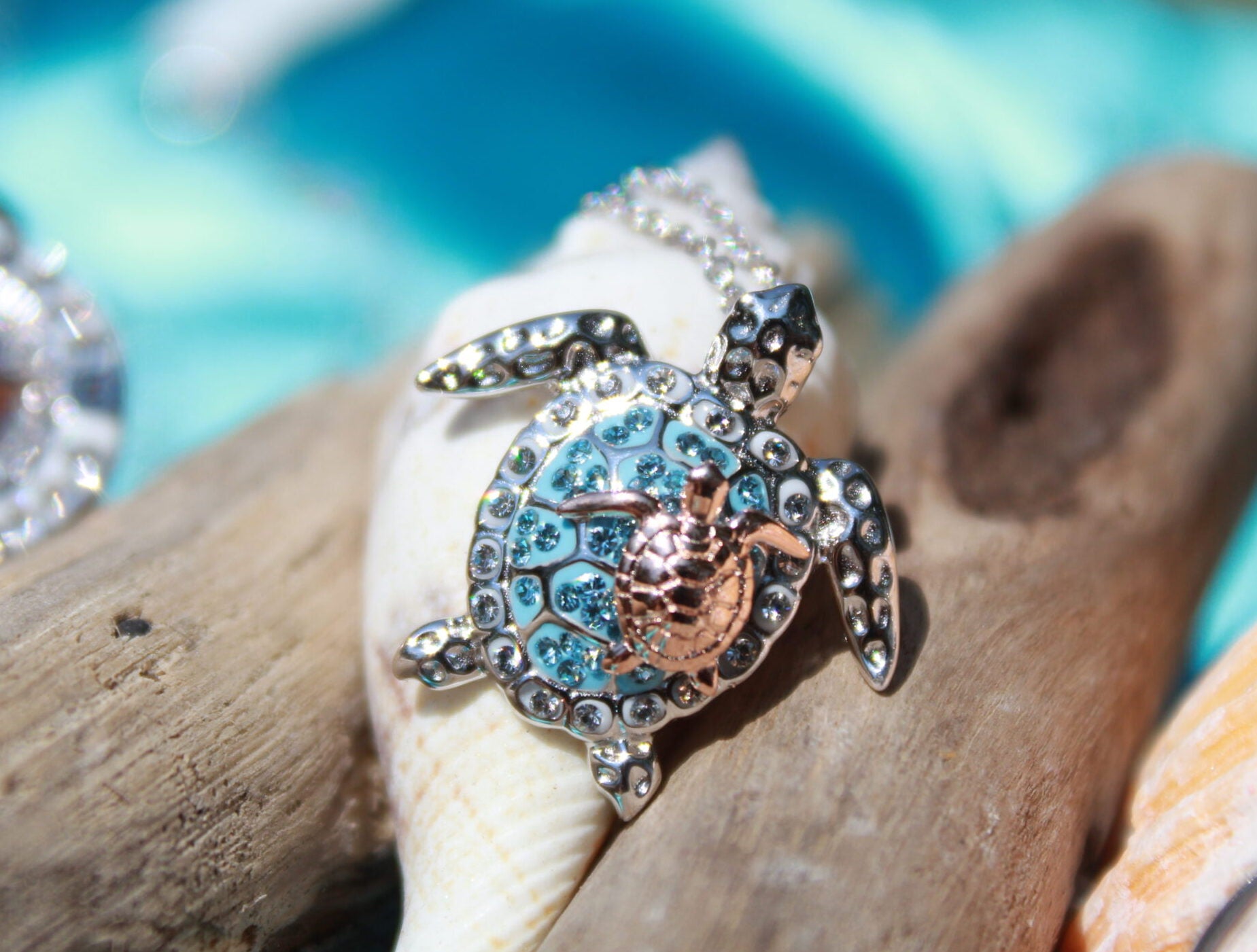 The Shanore Swarovski Crystals Aqua Turtle and Baby Necklace-Ocean Jewelry, crafted in sterling silver, features a turtle-shaped pendant with a blue gemstone-studded shell. It rests delicately on driftwood against a softly blurred beach backdrop of blue and sandy tones.