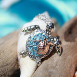 The Shanore Swarovski Crystals Aqua Turtle and Baby Necklace-Ocean Jewelry, crafted in sterling silver, features a turtle-shaped pendant with a blue gemstone-studded shell. It rests delicately on driftwood against a softly blurred beach backdrop of blue and sandy tones.