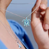A close-up shows someone wearing Shanores Swarovski Crystals March Birthstone Turtle Necklace, featuring a detailed turtle pendant with small blue stones. The background is a blurred sea and sky, and the person is in a blue top.