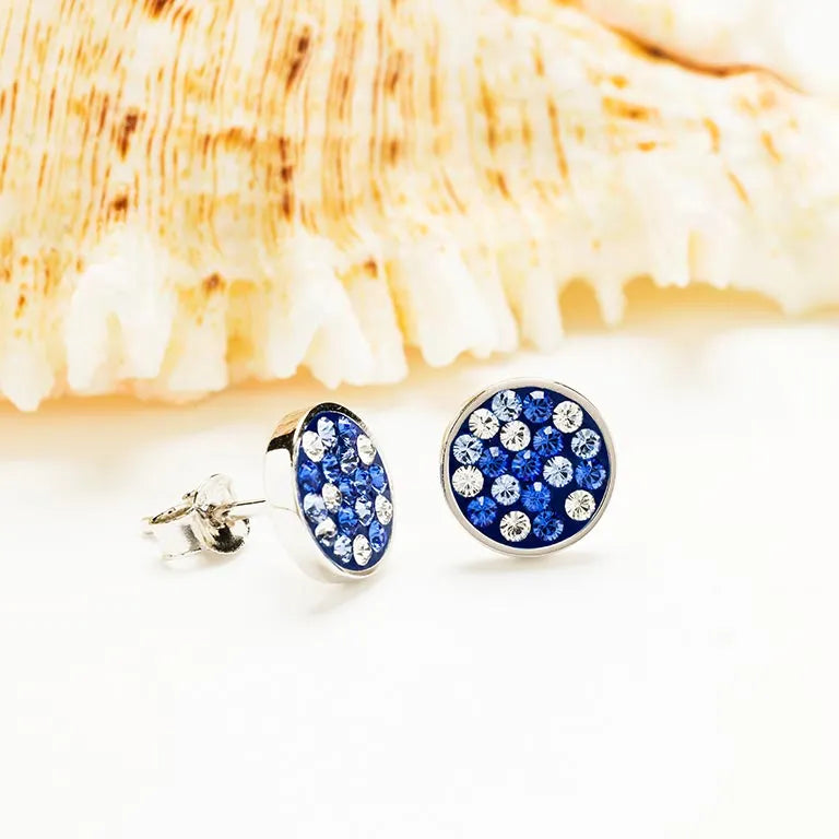 The Shanore Swarovski Crystals Blue Sapphire Round Stud Earrings elegantly showcase their beauty against a white backdrop, complemented by a textured seashell. These Ocean Jewelry earrings feature blue and white gemstones for added sparkle.