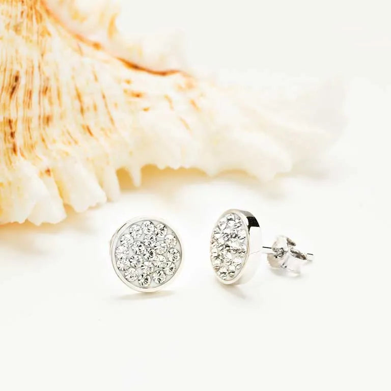 Swarovski Crystals White Stud Earrings- Ocean Jewelry by Shanore are elegantly placed on a soft white backdrop, complemented by a large cream seashell with orange accents.