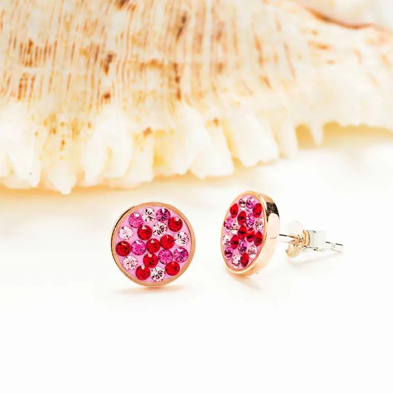 These Swarovski Crystals Pink Rose Stud Earrings by Shanore boast dazzling red and pink gemstones set in gold, elegantly displayed on a soft white seashell backdrop.