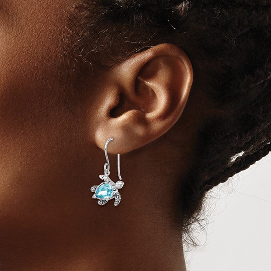 A close-up of a persons ear wearing Presco/Quality Gold Crystal Medium Blue Turtle Earrings, featuring a shiny blue gemstone shell with sterling silver head, legs, and tail.