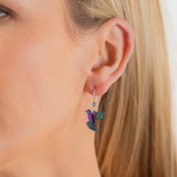 Close-up of a person with long blonde hair wearing Holly Yashis Island Green Picaflor Earrings, showcasing a colorful hummingbird design. The purple bird with teal wings is intricately hand-painted on niobium and hangs elegantly from sterling silver French wires against a white background.
