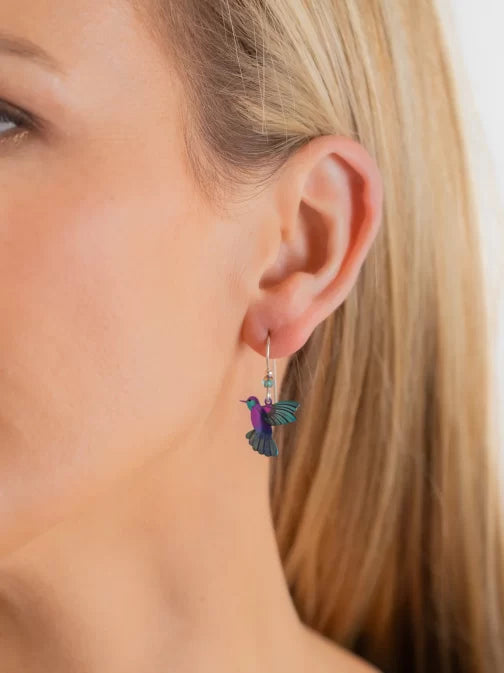 Close-up of a person with long blonde hair wearing Holly Yashis Island Green Picaflor Earrings, showcasing a colorful hummingbird design. The purple bird with teal wings is intricately hand-painted on niobium and hangs elegantly from sterling silver French wires against a white background.