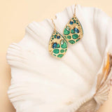 The Seashore Dreams Teal Earrings by Holly Yashi Jewelry elegantly rest on a white seashell against a beige backdrop. Their leaf-like shape with intricate detailing features vibrant blue-green gemstones and Austrian crystals, adding an extra touch of elegance.