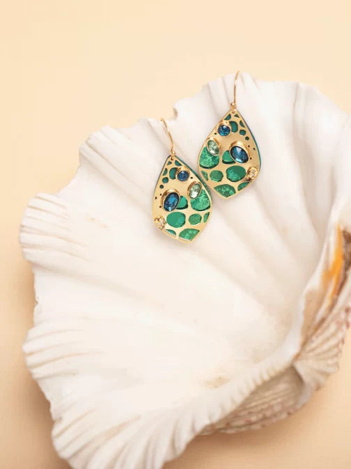 The Seashore Dreams Teal Earrings by Holly Yashi Jewelry elegantly rest on a white seashell against a beige backdrop. Their leaf-like shape with intricate detailing features vibrant blue-green gemstones and Austrian crystals, adding an extra touch of elegance.