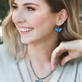 The woman smiles, flaunting Holly Yashis Green Flash Bella Butterfly Earrings and a matching necklace. Her shoulder-length blonde hair complements her light-colored top as her butterfly-shaped niobium earrings add whimsy to the blurred outdoor scene.