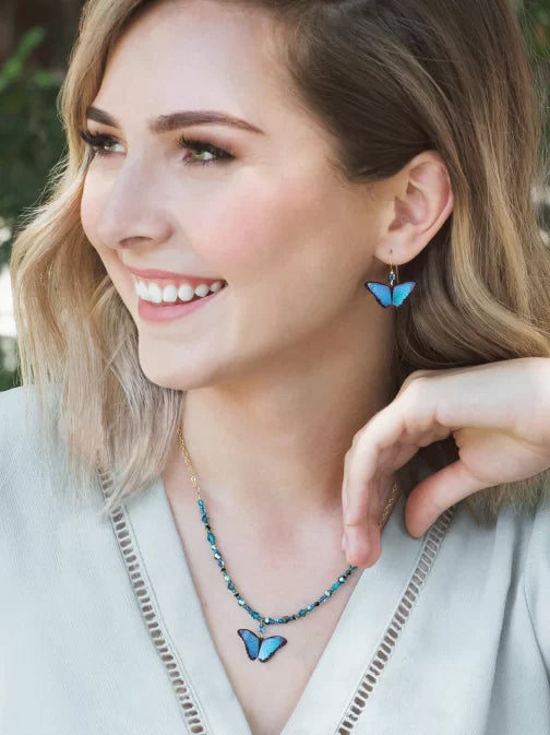 The woman smiles, flaunting Holly Yashis Green Flash Bella Butterfly Earrings and a matching necklace. Her shoulder-length blonde hair complements her light-colored top as her butterfly-shaped niobium earrings add whimsy to the blurred outdoor scene.