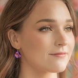 A close-up shows a woman with long brown hair wearing Holly Yashis Sparkling Fuchsia Garden Pansy Drop Earrings. With subtle makeup, she looks slightly to the side as colorful patterns blur softly in the background.
